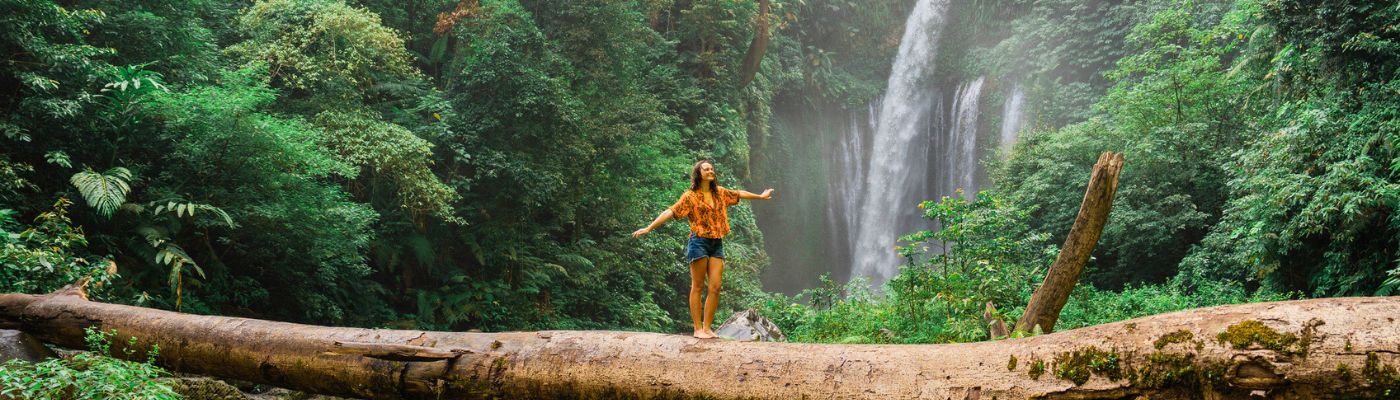 Woman crossing river by log on the background of tropical waterfall while hiking