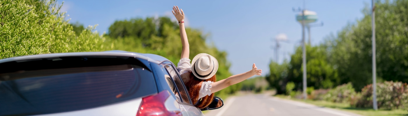 Happy young woman enjoying a summer road trip with arms out of car window, feeling free and joyful while traveling on a bright sunny day.