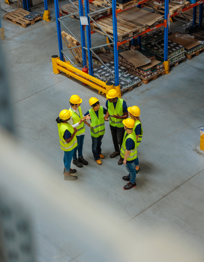 Group of warehouse workers in safety vests and helmets discussing in a large storage facility